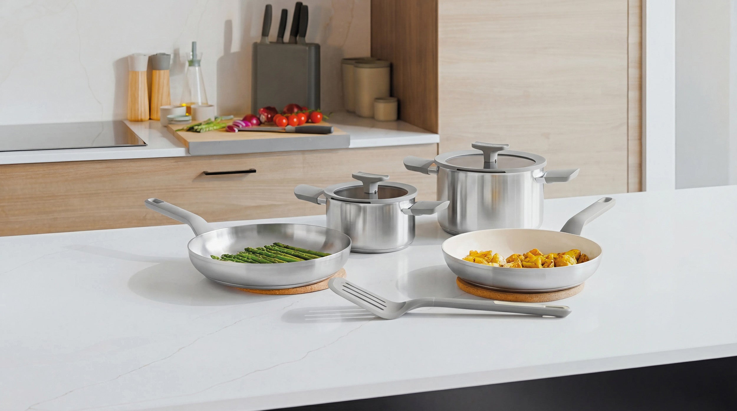 Stainless steel cookware set on a white countertop, including a stockpot, frying pan, and saucepans with wooden trivets.
