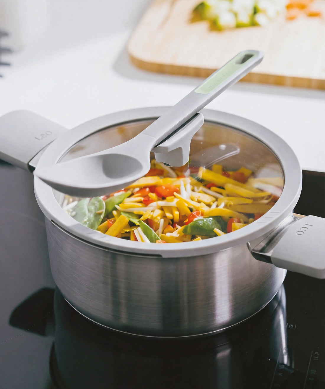 Stainless steel stockpot with a glass lid, filled with colorful chopped vegetables, on a stovetop.